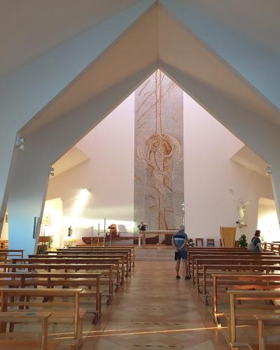 The interior of a modern church with white walls, wooden pews, and a large mosaic depicting the Assumption of Mary at the altar.