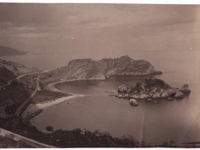 A sepia-toned vintage photograph showing a coastal landscape featuring a prominent limestone island connected to the mainland by a narrow sandy isthmus, with a road and small tunnel visible on the left side leading towards the landmass.