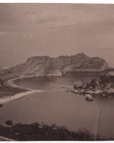 A sepia-toned vintage photograph showing a coastal landscape featuring a prominent limestone island connected to the mainland by a narrow sandy isthmus, with a road and small tunnel visible on the left side leading towards the landmass.