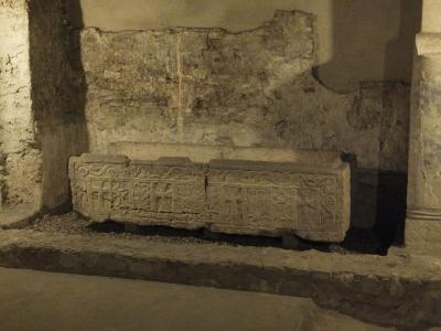 A stone sarcophagus with carved crosses and decorative motifs, displayed indoors against a rough, aged stone wall within an archaeological or museum setting.
