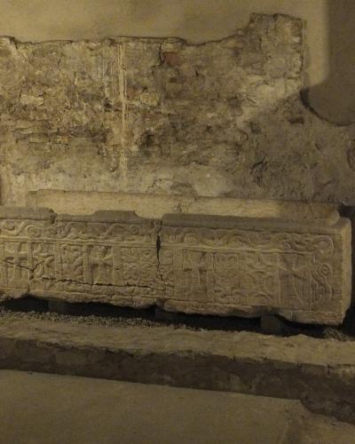A stone sarcophagus with carved crosses and decorative motifs, displayed indoors against a rough, aged stone wall within an archaeological or museum setting.