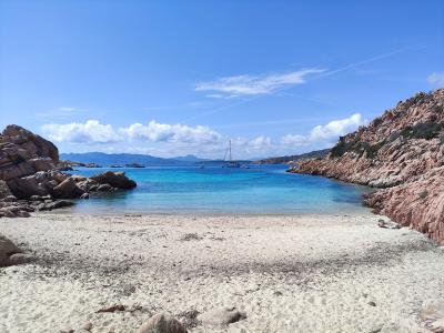 A scenic coastal view of a sandy beach framed by large pink granite boulders and rocky cliffs, with turquoise water, anchored boats, and distant mountains under a blue sky.