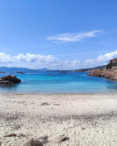 A scenic coastal view of a sandy beach framed by large pink granite boulders and rocky cliffs, with turquoise water, anchored boats, and distant mountains under a blue sky.