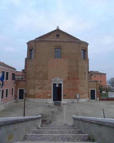 A large, cylindrical brick church with a triangular pediment and central entrance, viewed from a stone staircase leading up to a paved square, with adjacent buildings and a cloudy sky.