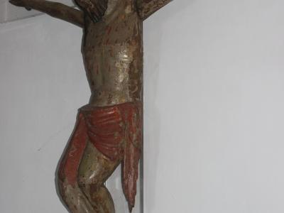 A weathered wooden crucifix of Jesus Christ mounted on a white wall, with visible paint wear and red accents, and part of a gold-framed object in the lower-left corner.