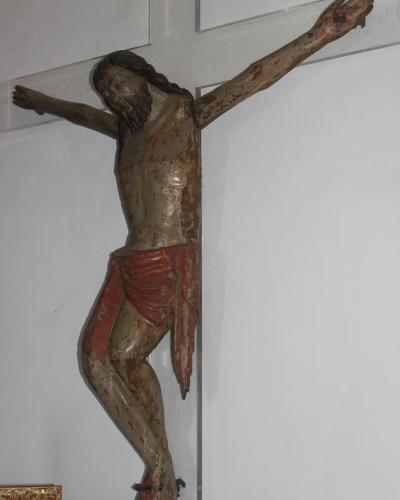 A weathered wooden crucifix of Jesus Christ mounted on a white wall, with visible paint wear and red accents, and part of a gold-framed object in the lower-left corner.
