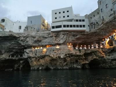 A view from the water looking up at a restaurant built into a limestone sea cave, with diners seated at tables under lights on a terrace, and white buildings perched above the cliff edge.