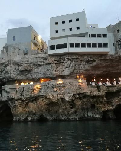 A view from the water looking up at a restaurant built into a limestone sea cave, with diners seated at tables under lights on a terrace, and white buildings perched above the cliff edge.