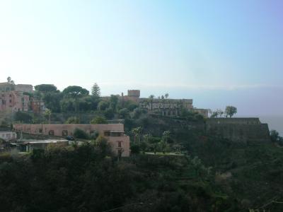 A long, terraced stone building with crenellated battlements and a central tower, situated on a steep, vegetated hillside overlooking the sea, with other residential buildings nearby.