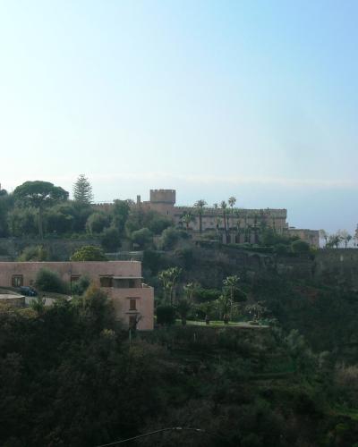 A long, terraced stone building with crenellated battlements and a central tower, situated on a steep, vegetated hillside overlooking the sea, with other residential buildings nearby.
