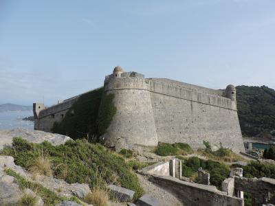 A large stone fortress with bastions and ivy-covered walls situated on a coastal hill overlooking the sea, with a small harbor and boats visible in the background.