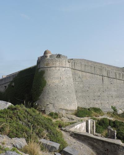 A large stone fortress with bastions and ivy-covered walls situated on a coastal hill overlooking the sea, with a small harbor and boats visible in the background.
