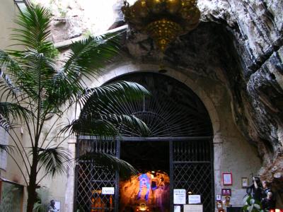 The image shows the entrance to a grotto-style sanctuary with a large golden candelabra hanging above an arched metal gate, flanked by potted palm trees and votive candles, with signage reading "ENTRATA" visible on the gate.
