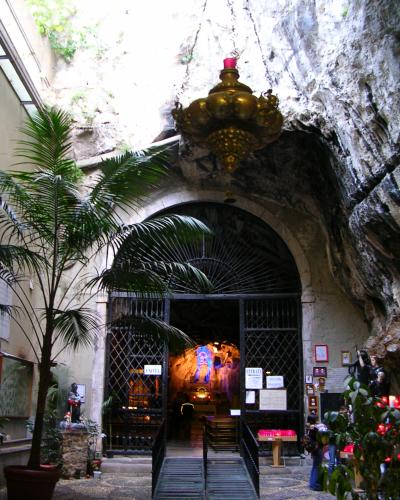The image shows the entrance to a grotto-style sanctuary with a large golden candelabra hanging above an arched metal gate, flanked by potted palm trees and votive candles, with signage reading "ENTRATA" visible on the gate.