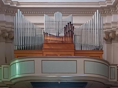 A close-up interior view of a large pipe organ with silver and wooden pipes, mounted on a curved balcony within a building featuring classical architectural details like pilasters and cornices.
