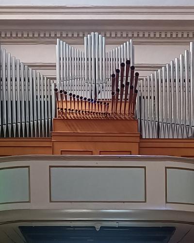 A close-up interior view of a large pipe organ with silver and wooden pipes, mounted on a curved balcony within a building featuring classical architectural details like pilasters and cornices.