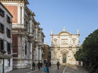 The image shows a sunlit Venetian square (campo) with a large, ornate white marble church facade on the right and a long, classical building with pilasters and red brickwork on the left; pedestrians walk across the paved plaza under a clear blue s