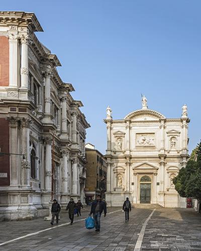 The image shows a sunlit Venetian square (campo) with a large, ornate white marble church facade on the right and a long, classical building with pilasters and red brickwork on the left; pedestrians walk across the paved plaza under a clear blue s