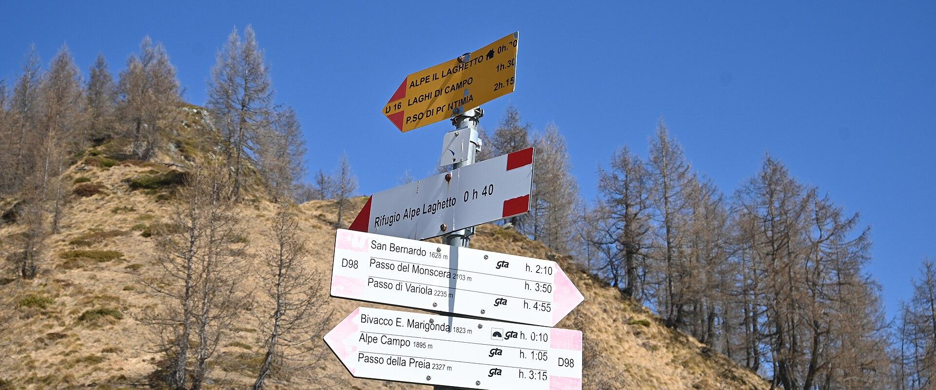 A trail signpost in a mountainous, alpine environment with dry grass and larch trees under a clear blue sky; signs point to locations including "Alpe il Laghetto," "Rifugio Alpe Laghetto," "Passo del Monscero," and "Bivacco E. Mangonda," with hiki