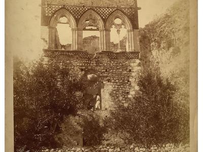 A sepia-toned photograph of a stone structure featuring a Gothic-style loggia with three pointed arches and crenellations, set against a hillside with a low stone wall and vegetation in the foreground.