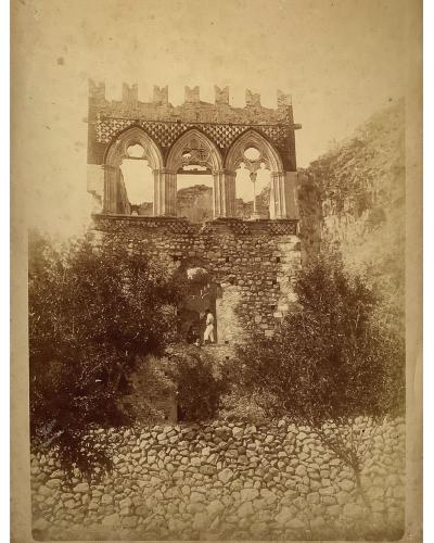 A sepia-toned photograph of a stone structure featuring a Gothic-style loggia with three pointed arches and crenellations, set against a hillside with a low stone wall and vegetation in the foreground.