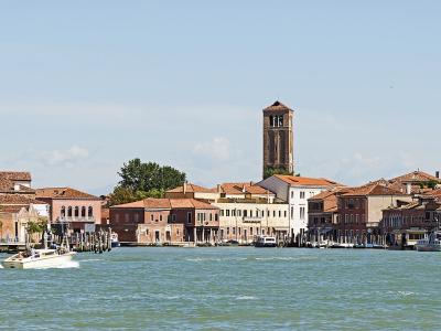 A panoramic view of a Venetian lagoon island featuring a prominent brick bell tower, terracotta-roofed buildings along the waterfront, and vaporetti (water buses) on the canal.