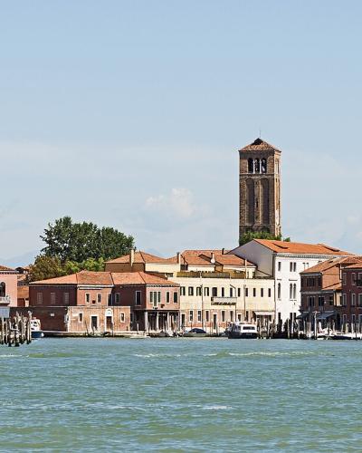 A panoramic view of a Venetian lagoon island featuring a prominent brick bell tower, terracotta-roofed buildings along the waterfront, and vaporetti (water buses) on the canal.