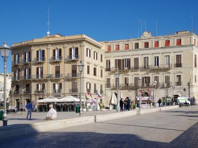 A sunlit, wide paved public square in an Italian city, featuring large beige historic buildings with balconies and shuttered windows, outdoor café seating under umbrellas, pedestrians, and a clear blue sky.