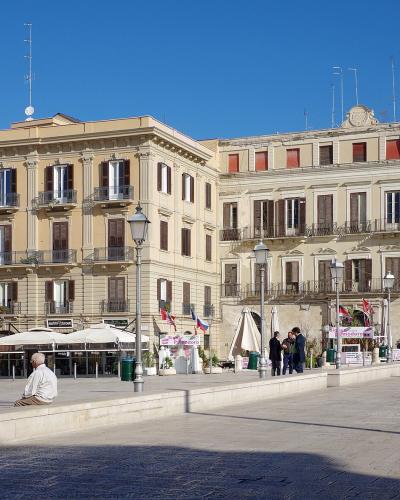 A sunlit, wide paved public square in an Italian city, featuring large beige historic buildings with balconies and shuttered windows, outdoor café seating under umbrellas, pedestrians, and a clear blue sky.