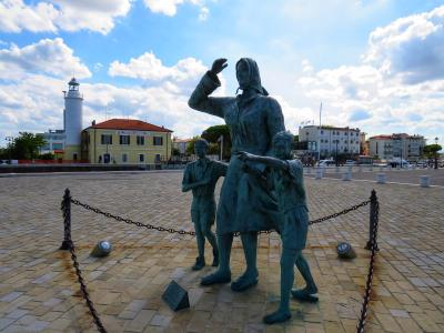 A bronze statue of a woman and two children in the foreground, with the white Cesenatico Lighthouse visible in the background on the left, set against a blue sky with clouds over a paved plaza.