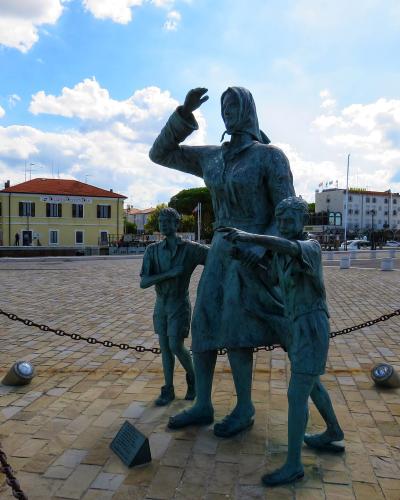 A bronze statue of a woman and two children in the foreground, with the white Cesenatico Lighthouse visible in the background on the left, set against a blue sky with clouds over a paved plaza.