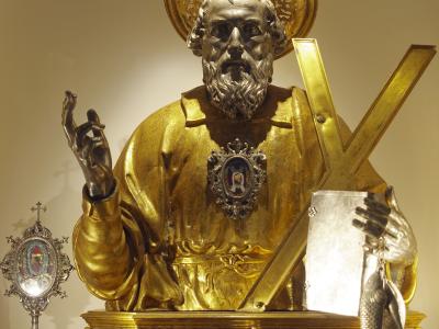A gilded and silvered bust statue of Saint Andrew holding an X-shaped cross and two fish, displayed on an ornate silver and gold reliquary base with a small plaque reading "S. ANDREA" and a silver monstrance to the left.