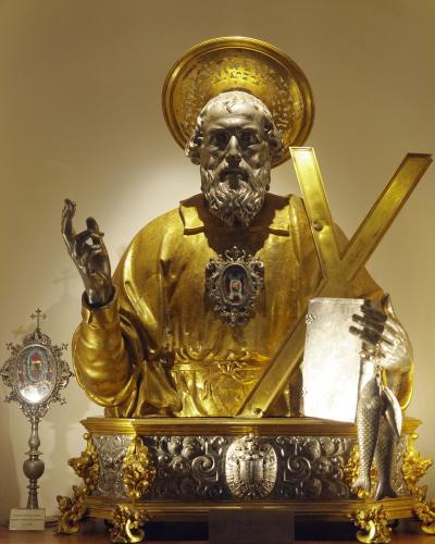 A gilded and silvered bust statue of Saint Andrew holding an X-shaped cross and two fish, displayed on an ornate silver and gold reliquary base with a small plaque reading "S. ANDREA" and a silver monstrance to the left.
