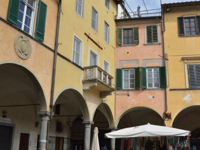 A sunlit Italian piazza with yellow and peach-colored buildings featuring green shutters, stone arches, and a white market tent set up on the cobblestone ground.