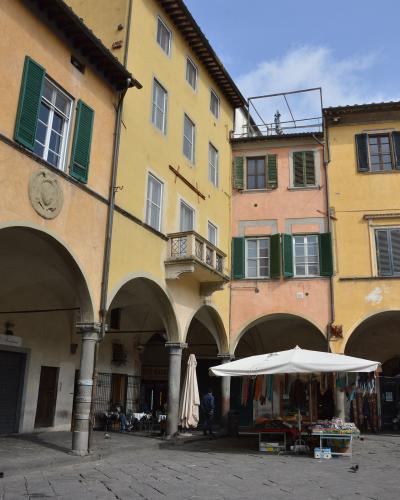 A sunlit Italian piazza with yellow and peach-colored buildings featuring green shutters, stone arches, and a white market tent set up on the cobblestone ground.