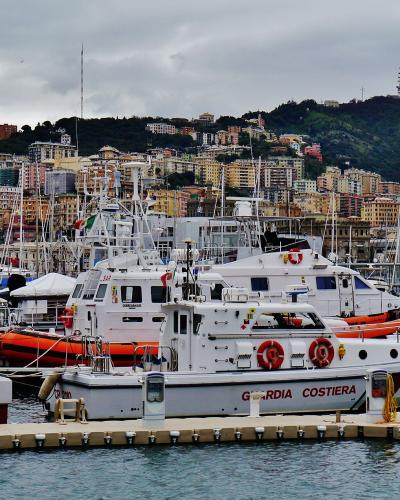 The image shows a harbor with several white Coast Guard (Guardia Costiera) boats docked at a pier, with a densely built hillside city in the background under an overcast sky.