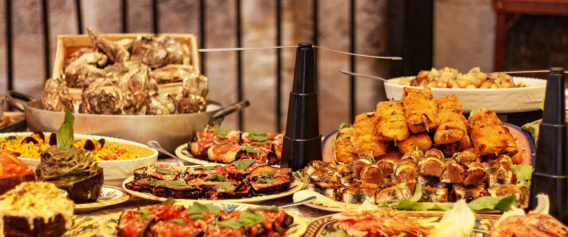 A table laden with various Italian dishes including stuffed eggplants, fried foods, and seafood, set against a wooden backdrop with decorative tiles on the tablecloth.