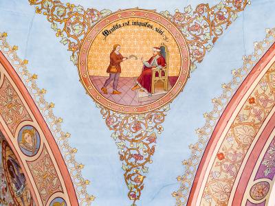 A close-up photograph of an ornate, late Baroque or Rococo-style painted ceiling vault featuring a central medallion with a religious scene and Latin text, surrounded by elaborate floral and scrollwork stucco or fresco decorations.