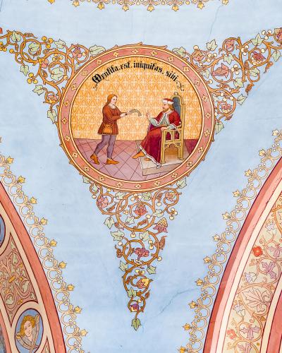 A close-up photograph of an ornate, late Baroque or Rococo-style painted ceiling vault featuring a central medallion with a religious scene and Latin text, surrounded by elaborate floral and scrollwork stucco or fresco decorations.