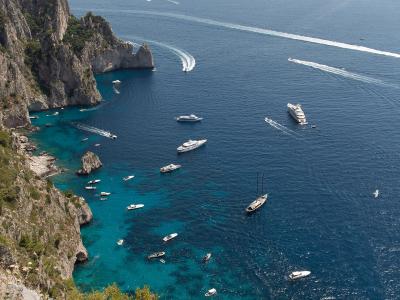 A high-angle scenic view of the coastline of Capri, Italy, showing numerous boats in the turquoise and deep blue waters near steep cliffs, with the island of Nisida visible in the distance.