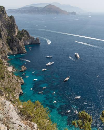 A high-angle scenic view of the coastline of Capri, Italy, showing numerous boats in the turquoise and deep blue waters near steep cliffs, with the island of Nisida visible in the distance.