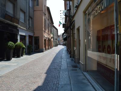 A sunlit, narrow European street paved with cobblestones and flanked by multi-story buildings housing retail shops, including a "BRIAN BARRY" store on the left and a "motivi" store on the right, with an Italian flag hanging from a building further