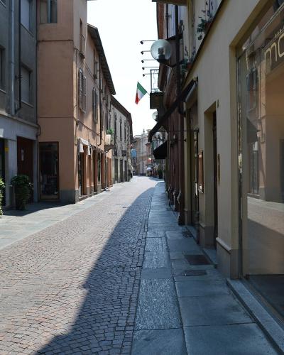 A sunlit, narrow European street paved with cobblestones and flanked by multi-story buildings housing retail shops, including a "BRIAN BARRY" store on the left and a "motivi" store on the right, with an Italian flag hanging from a building further