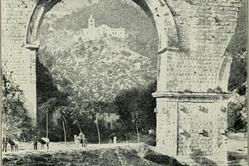 A black-and-white photograph showing a large, ancient stone archway (likely part of a Roman aqueduct or bridge) with a hillside village and church visible through the arch; people and donkeys are present at the base.