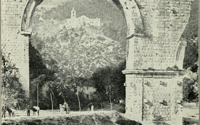 A black-and-white photograph showing a large, ancient stone archway (likely part of a Roman aqueduct or bridge) with a hillside village and church visible through the arch; people and donkeys are present at the base.