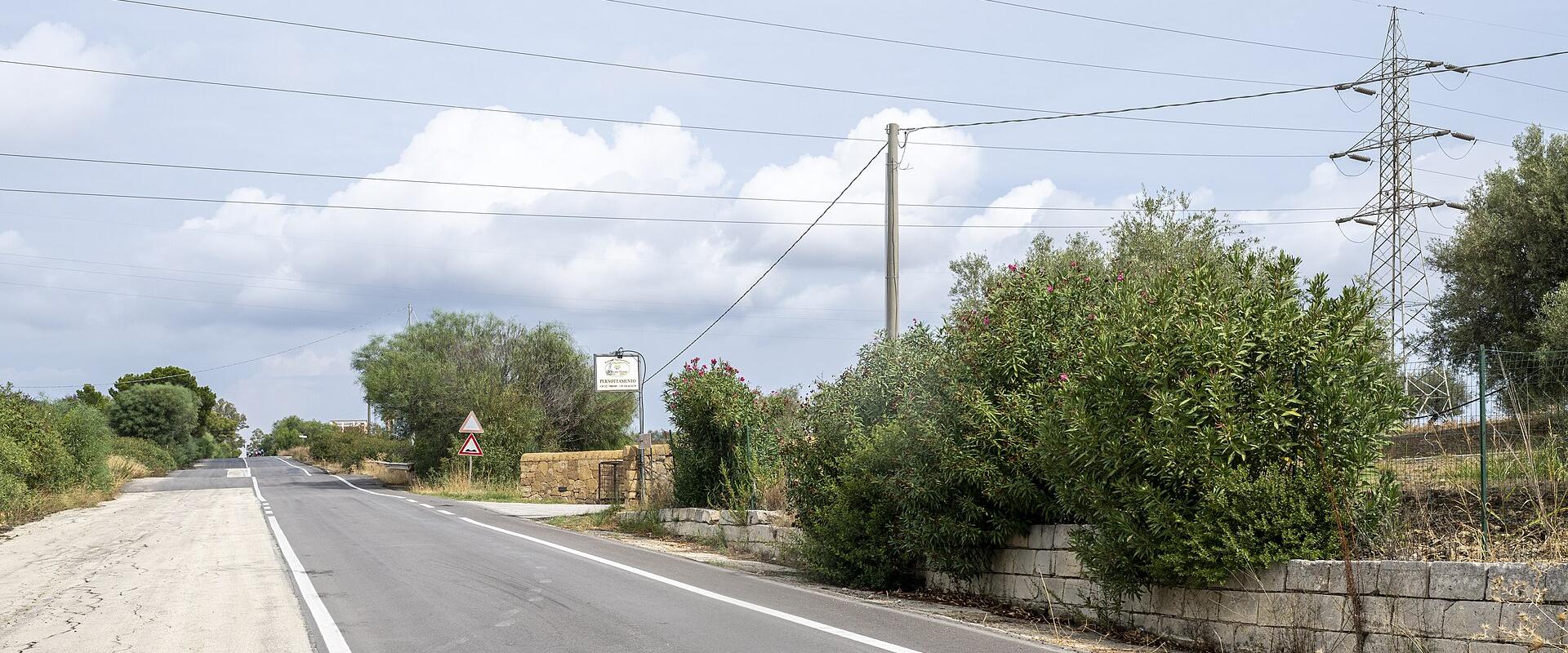 A paved two-lane road in a rural Mediterranean landscape with olive trees, oleander bushes, stone walls, overhead power lines, and a triangular warning sign; a white rectangular signpost is visible near the roadside but its text is illegible.