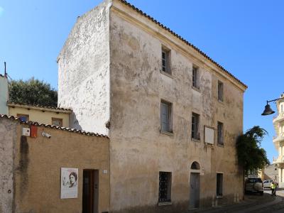 A weathered, multi-story stucco building on a street corner with a poster of Grazia Deledda on an adjacent wall and a commemorative plaque on the facade.