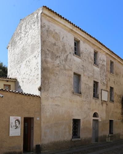 A weathered, multi-story stucco building on a street corner with a poster of Grazia Deledda on an adjacent wall and a commemorative plaque on the facade.