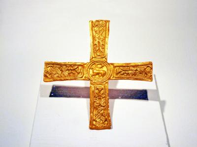 A close-up photograph of an ornate gold cross with intricate animal and geometric engravings, displayed on a white museum stand against a plain background.