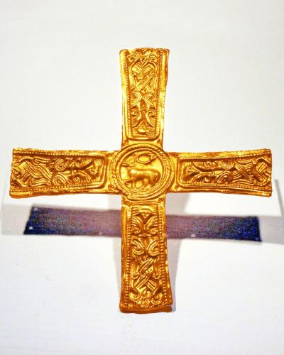 A close-up photograph of an ornate gold cross with intricate animal and geometric engravings, displayed on a white museum stand against a plain background.